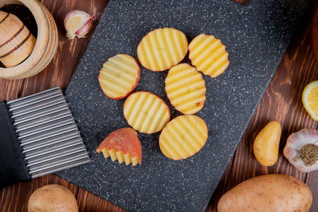 top view of ruffled slices of potatoes on cutting board with whole ones lemon garlic around on wooden backgroundの写真素材