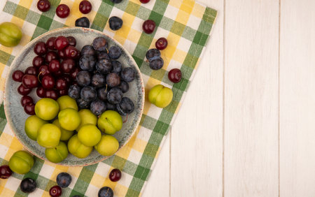 top view of fresh fruits such as cherriessloes and green cherry plums on a bowl on a checked tablecloth on a white background with copy spaceの写真素材