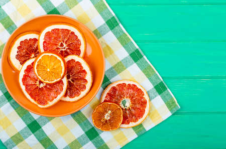 top view of dried orange and grapefruit slices in a plate on plaid tablecloth on green wooden background with copy spaceの写真素材