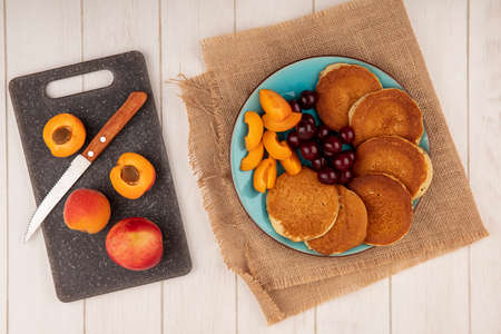top view of pancakes with cherries and apricot pieces in plate on sackcloth and apricots with knife on cutting board on wooden backgroundの写真素材