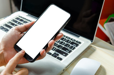 Man hand is pressing the smartphone with a blank screen for designing or advertising on the work desk and the laptop computer on the work table.の素材