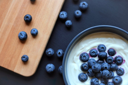 A close-up overhead view shows fresh blueberries scattered on a light wooden cutting board and piled...