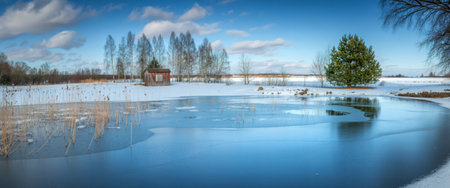 A serene winter scene featuring a partially frozen pond reflecting the sky, surrounded by snow-covered ground and bare trees, with a small gazebo in the distance.の写真素材
