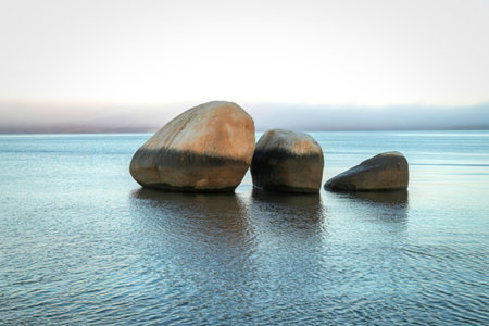 Three large, smooth, rounded rocks stand in shallow, reflective water. The sky above is a soft, hazy gradient.の写真素材