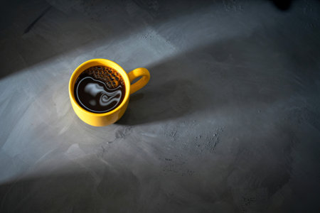 A bright yellow mug filled with steaming black coffee sits on a textured gray surface, illuminated by a dramatic shaft of light.の写真素材