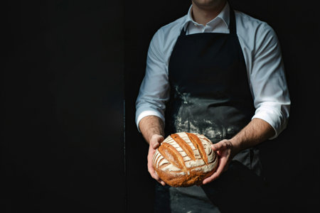 A baker in a white shirt and black apron holds a beautifully scored, golden-brown sourdough loaf against a dark background.の写真素材