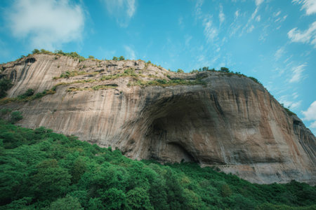 A dramatic, textured cliff face rises from dense green foliage, reaching towards a vast sky filled...