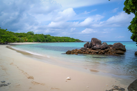 A serene tropical beach with calm turquoise waters, white sand, and a cluster of dark rocks. Lush green trees line the shore under a cloudy sky.の写真素材