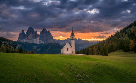 A solitary white church stands in a green meadow with the dramatic peaks of the Dolomites and a vibrant sunset sky.の写真素材