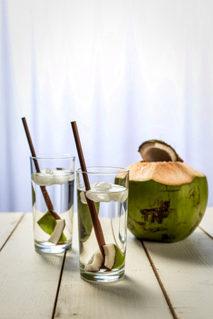 Two glasses of clear liquid with lime and straws sit next to a halved green coconut on a white wooden surface.の写真素材