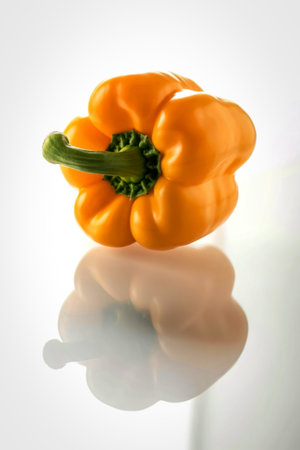 A vibrant yellow bell pepper with a green stem sits on a reflective white surface, casting a soft...