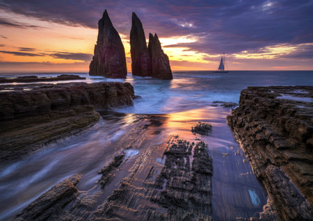 Jagged rock spires rise from the sea as waves crash over rocky shores during a vibrant sunset.
