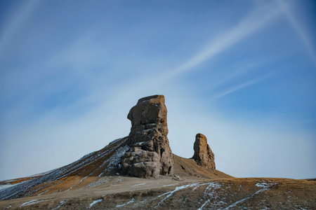A striking, weathered rock spire dominates the landscape under a bright, expansive sky with faint...