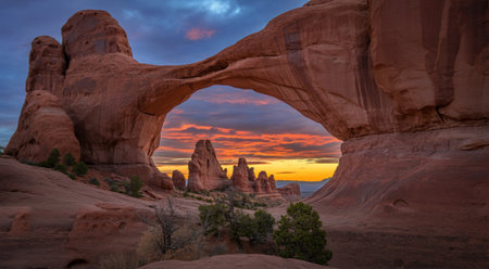 A vibrant sunset paints the sky with fiery colors, viewed through a massive natural sandstone arch,...