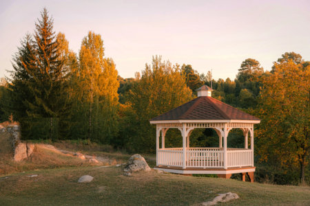 A charming white gazebo stands amidst a vibrant autumn forest as the sun sets, casting a warm glow.の写真素材