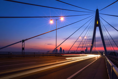 A striking cable-stayed bridge at twilight, showcasing the city skyline and streaking car lights on the roadway below.の写真素材