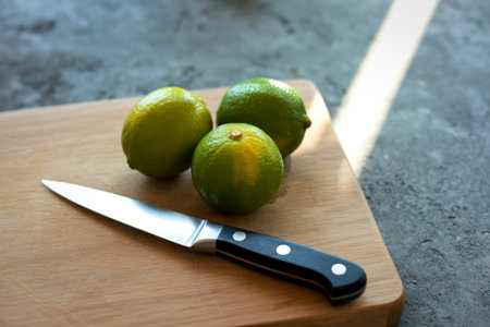 Three vibrant green limes sit on a wooden cutting board next to a sharp kitchen knife, ready for preparation.の写真素材