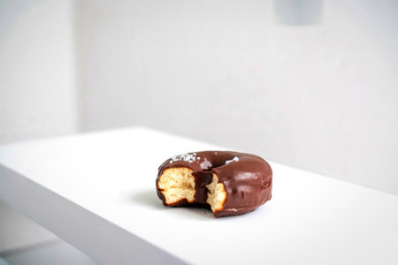 A close-up shot of a chocolate-glazed donut with a bite taken out, revealing its fluffy interior, placed on a white surface.の写真素材