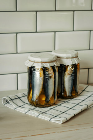 Two glass jars filled with preserved sardines in oil sit on a checkered cloth against a white subway tile background.の写真素材