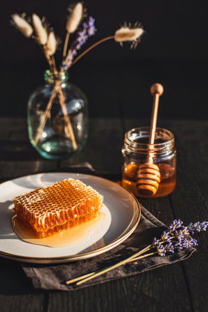 A piece of fresh honeycomb sits on a white plate next to a jar of honey with a wooden dipper and dried floral accents.の写真素材