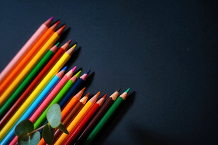 A collection of brightly colored pencils diagonally arranged on a dark surface, with a hint of greenery in the foreground.の写真素材