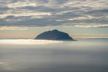 A solitary island peak rises majestically from a thick blanket of sea fog, with dramatic clouds...