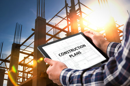 A person in a plaid shirt holds a tablet displaying construction plans against a backdrop of a...