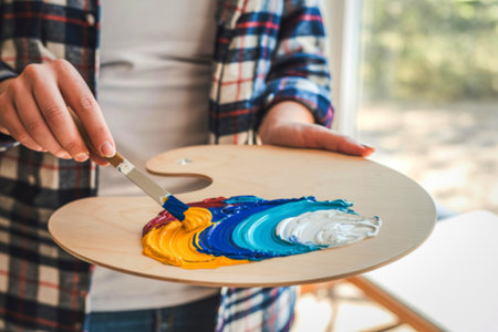 A person in a plaid shirt holds a wooden palette with vibrant oil paints being mixed by a palette knife.の写真素材