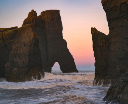 Towering rock formations rise from the ocean as waves crash against them during a soft pastel...