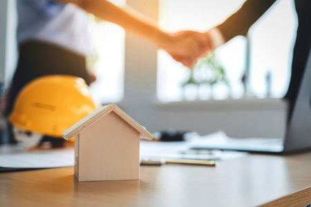 Two people shake hands over a table with a model house, hard hat, and blueprints, signifying a...