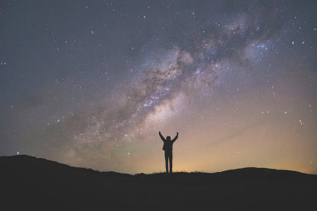A person stands with arms raised, silhouetted against the stunning backdrop of the Milky Way galaxy....
