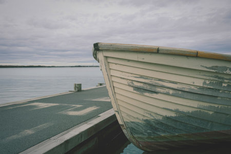 A weathered wooden boat rests on a dock beside tranquil waters under a cloudy sky.