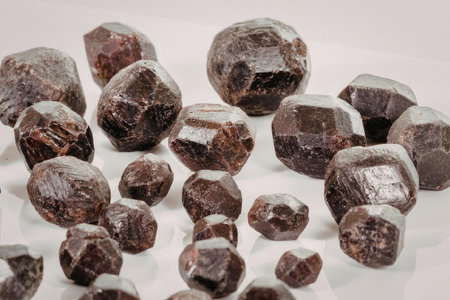 A close-up view of numerous dark red gemstones of various sizes scattered on a white surface,...