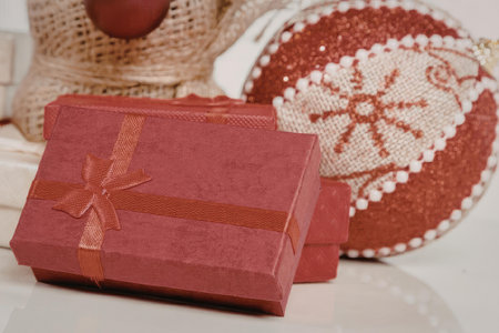 A red gift box with a ribbon, a red and white ornament, and a burlap object are placed on a white...