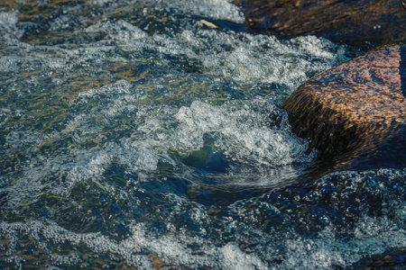 A close-up view of water flowing over a rock in a river or stream, creating white foam and bubbles.