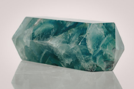 A green crystal with a unique shape sits on a white surface. The crystal has a mix of light and dark...
