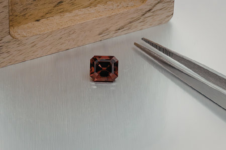 A small, square-cut red gemstone with a black center sits on a clear plastic stand next to a pair of...