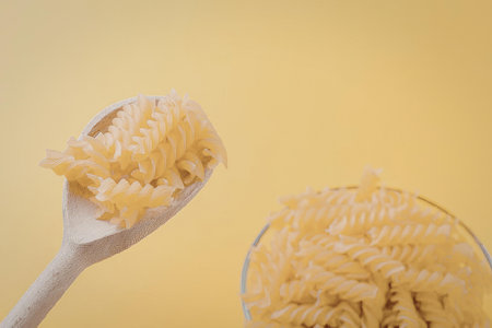 A wooden spoon holding fusilli pasta above a glass bowl filled with more fusilli pasta, set against...