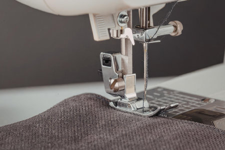A close-up view of a sewing machine's metal parts and needle as it sews through a dark gray fabric.