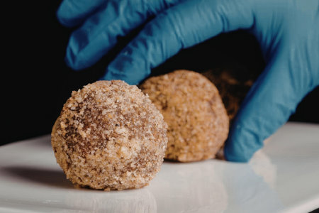 a close-up of a blue-gloved hand reaching for two fried balls coated in breadcrumbs on a white plate