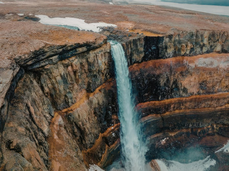 An aerial view of a breathtaking waterfall cascading down a rugged cliffside, surrounded by a vast, barren landscape with patches of snow and vibrant orange and green hues.の写真素材