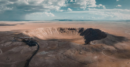 An aerial view of a vast desert landscape featuring a large crater with rugged terrain and a blue sky with clouds.の写真素材
