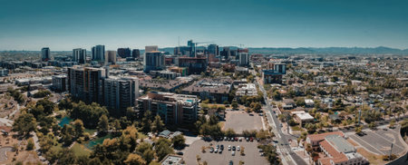 A panoramic view of a cityscape with tall buildings, trees, and a clear blue sky. The image...