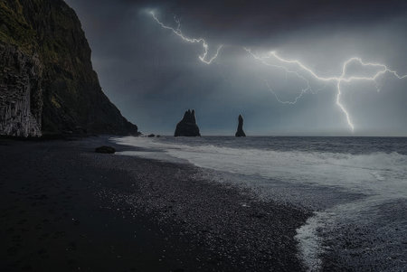A dark and moody landscape of a rocky coastline with waves crashing on the shore under a stormy sky with lightning.の写真素材