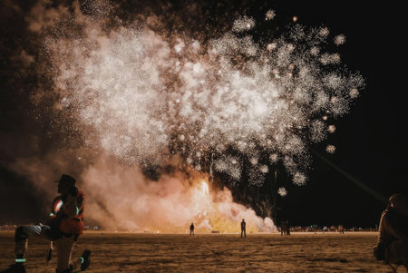 A nighttime scene of a large fire in a field with people standing around, fireworks exploding in the...
