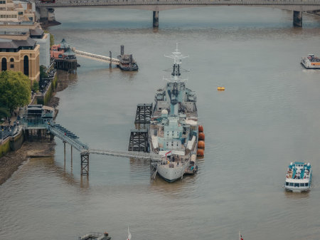 A large warship is docked at a pier on a river, with several smaller boats nearby and a bridge in the background.の写真素材