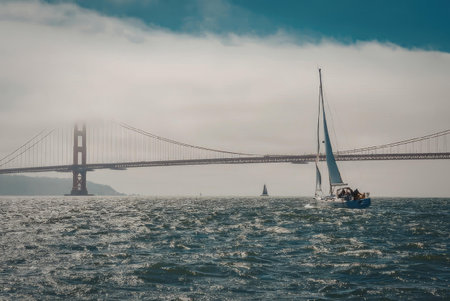 A sailboat sailing under the Golden Gate Bridge on a sunny day with a cloudy sky