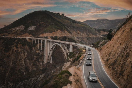 A picturesque highway with a large bridge curves through a mountainous landscape during sunset.