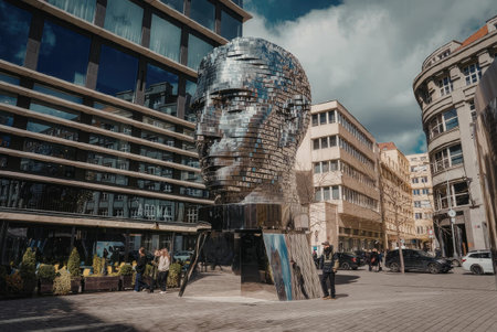 A large metal sculpture of a face is situated in a city square surrounded by modern and old...