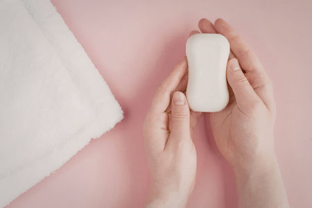 A pair of hands holding a white soap bar with a white towel on a pink background